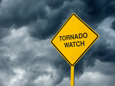 Diamond shaped yellow sign with black text reading tornado watch with stormy clouds in background.