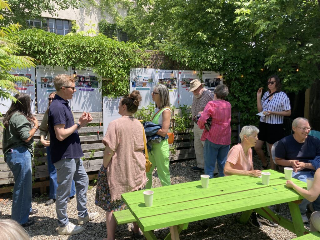 Members of the public gathering and casually chatting outside beside whiteboards showing community requests.