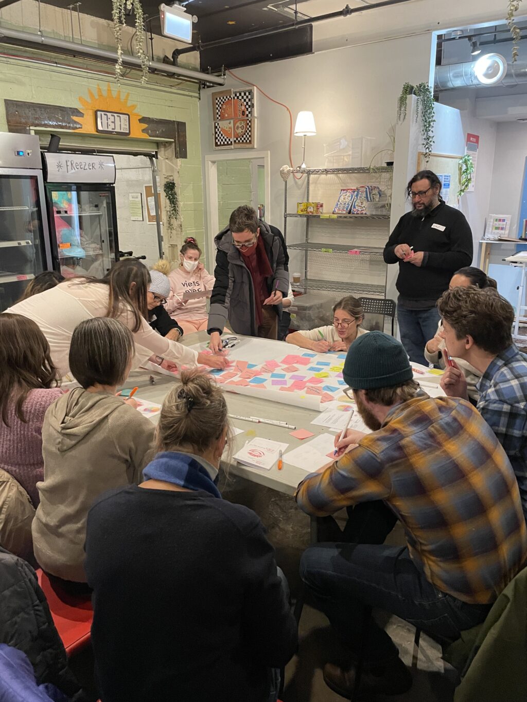 Community gathered around a table writing down responses to questions on sticky notes and sticking them on a whiteboard.