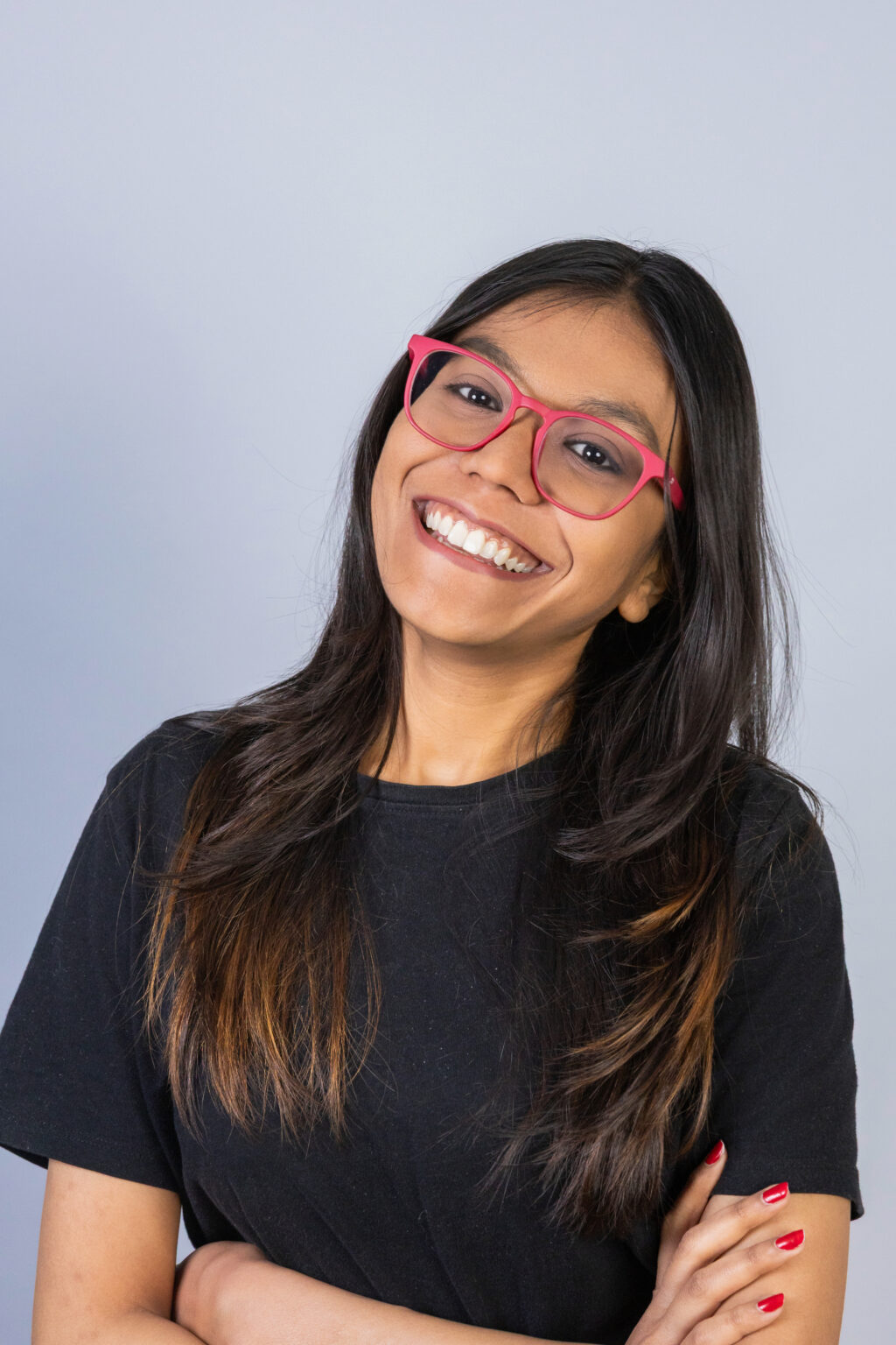 Devanshee Vankani headshot. Posing in front of a grey background. She is wearing a black t-shirt and pink glasses. 