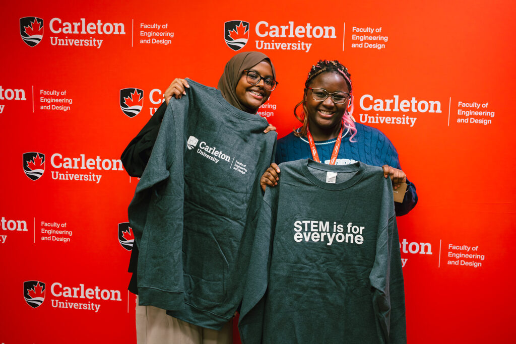 Two women students posing with sweaters that say "STEM is for everyone" with a red Carleton University backdrop. 