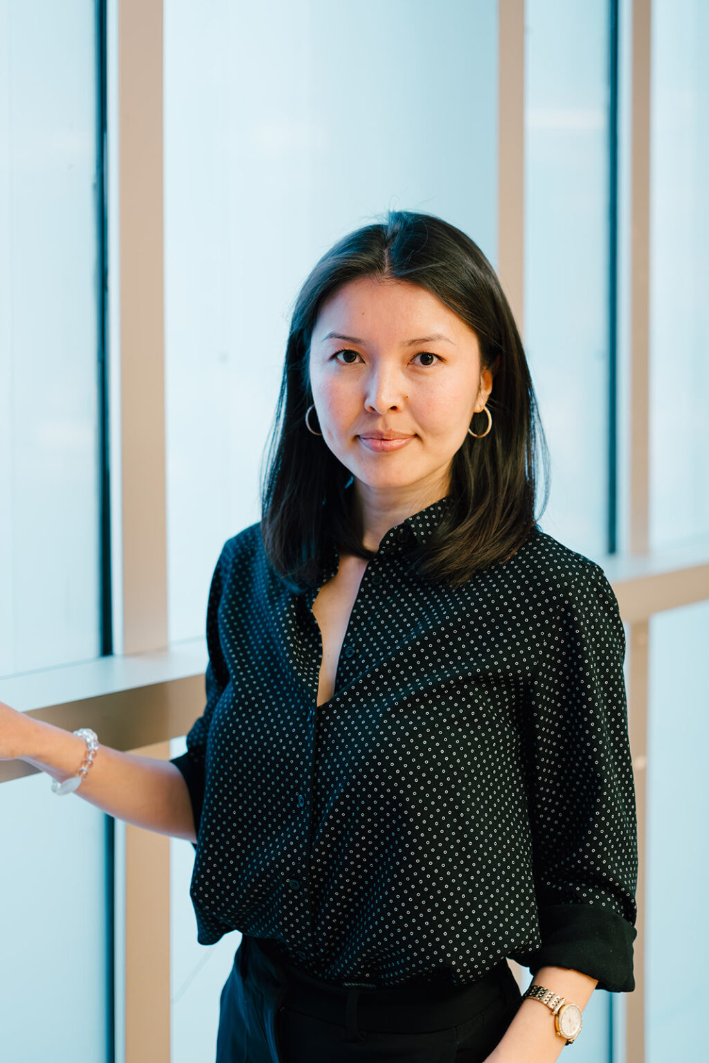 Portrait of Diana Tutkishbayeva, posing with windows in the back, wearing a black polka dot blouse. 