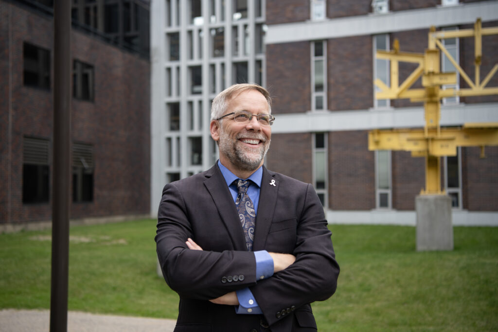 Ron Miller with his arms crossed, smiling and looking away from the camera. He's standing on campus by the Minto building