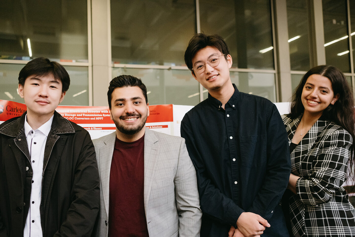 Three students are smiling and standing together indoors. They are dressed in smart casual attire, with the two on either side wearing blazers. The background features a display board with some text.