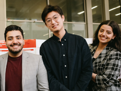 Three students are smiling and standing together indoors. They are dressed in smart casual attire, with the two on either side wearing blazers. The background features a display board with some text.