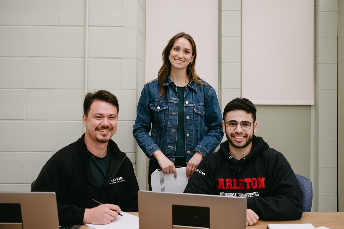Three people are smiling in a study or work setting. Two are seated at a table with laptops, papers, and pens, while one stands behind them.