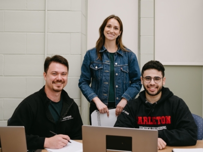 Three people are smiling in a study or work setting. Two are seated at a table with laptops, papers, and pens, while one stands behind them.