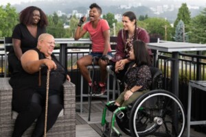 Five people talking. Two Black women, One white woman with a  cane, one Asian women in a wheelchair and another White woman sitting down. 