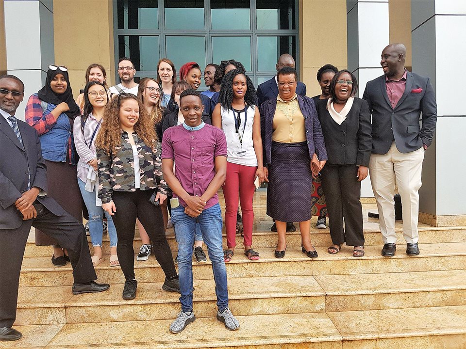 Group of people standing on outdoor steps in front of a building with large windows, posing casually for a group photo.