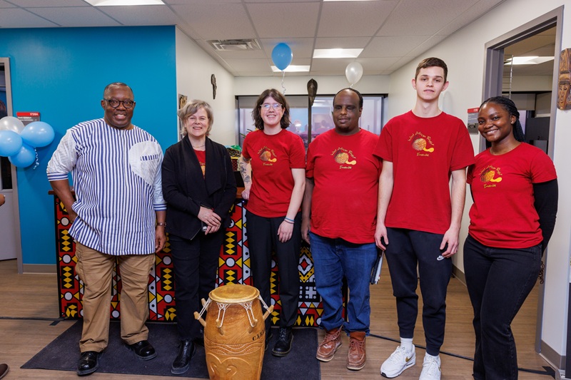 Five people stand indoors behind a large carved drum, with colorful African décor and balloons in the background.