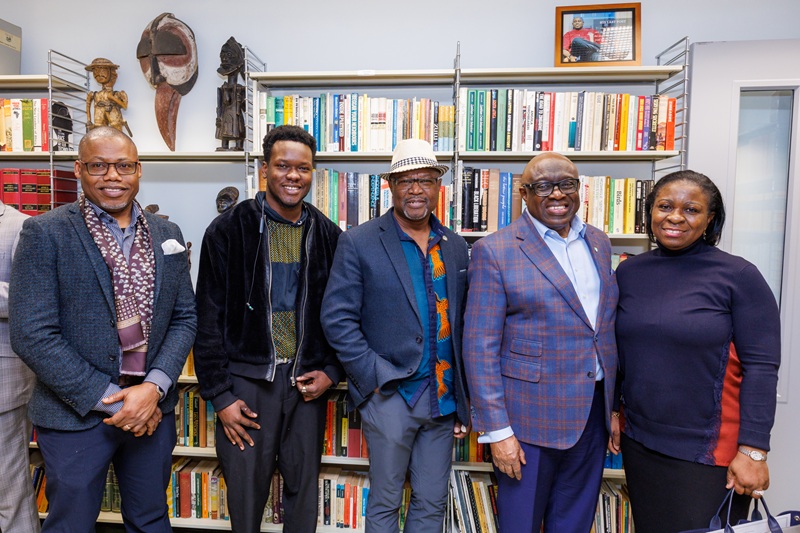 Five black people stand in front of a  bookshelf filled with books and African art, in a professional setting.