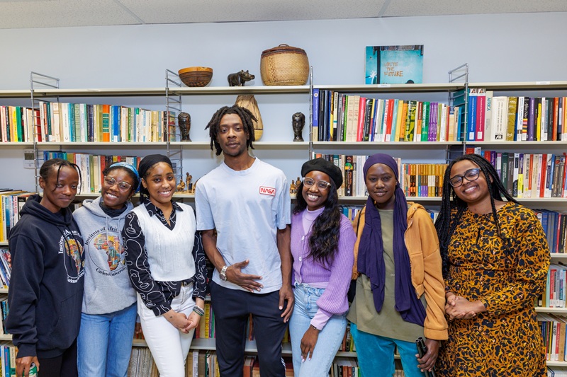 Seven Black students stand together in front of a large bookshelf filled with books and decorative items.
