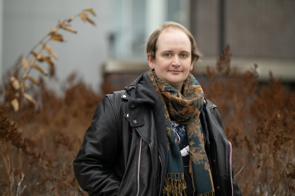 Simon standing outside in the fall. Simon has blue eyes and blonde hair, and is wearing a leather jacket and orange scarf.