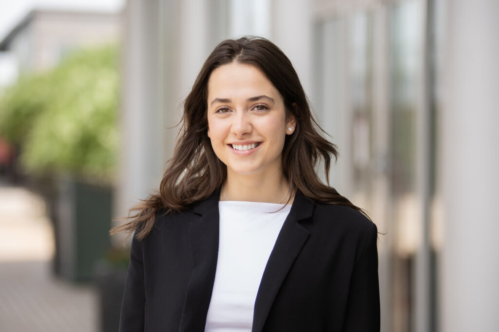 Ayla Sully has brown shoulder-length hair, light skin, and brown eyes, and is wearing a white top and black blazer. 