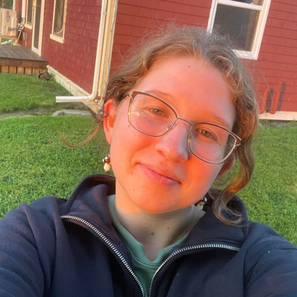 Student Ambassador in navy sweater with glasses, i front of red cottage