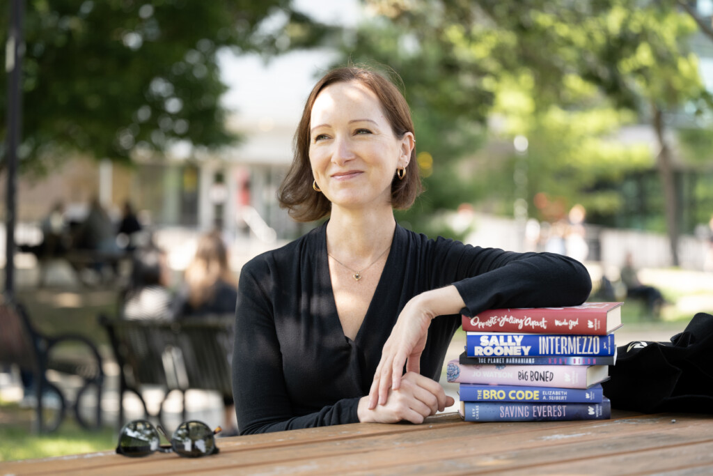 Professor Sarah Brouillette sitting on a bench in the Quad, resting her arm on a stack of books.