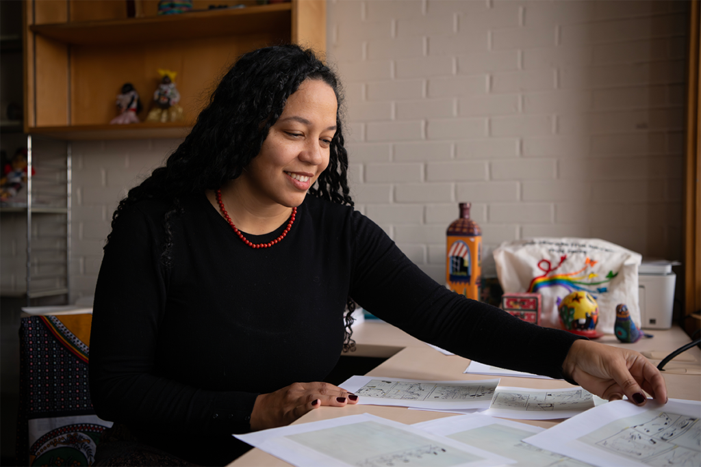 A person sits at a desk reviewing and arranging illustrated pages in an office workspace.