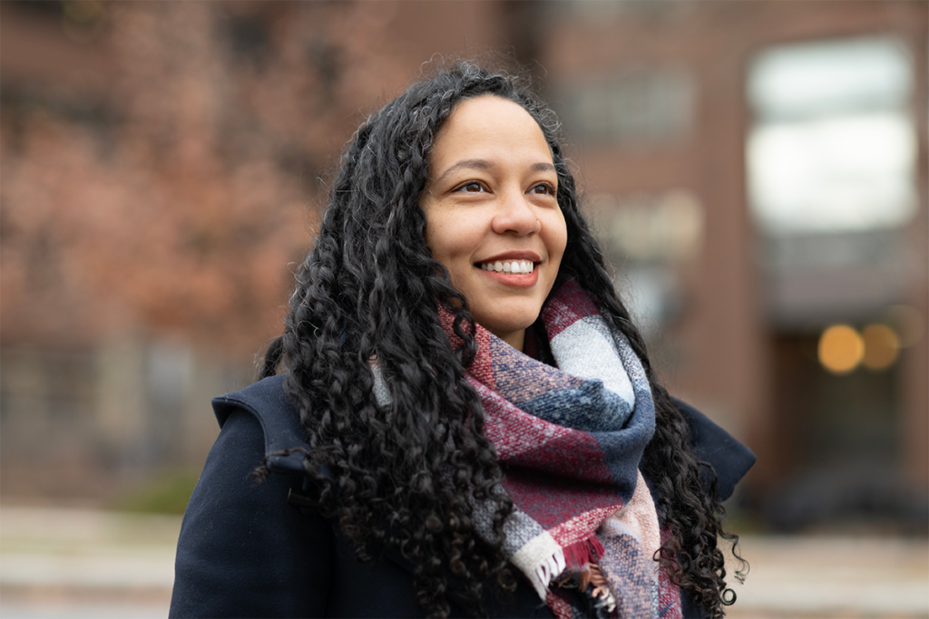 Close-up portrait of a person wearing a dark coat and plaid scarf outdoors, with a brick building in the background.