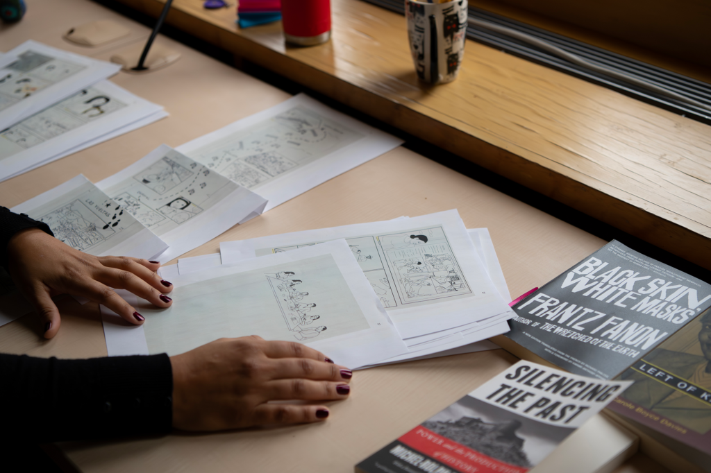 Hands sort through printed storyboard or comic pages on a desk beside several books and reference materials near a window.
