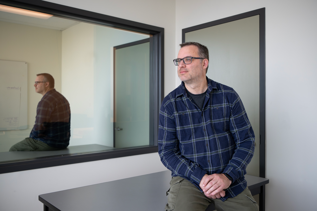 Person wearing glasses and a blue plaid shirt sits on a table in a bright office space.