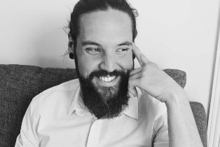Black-and-white portrait of a person with long hair tied back and a full beard, seated on a couch and resting one hand against the side of their face.