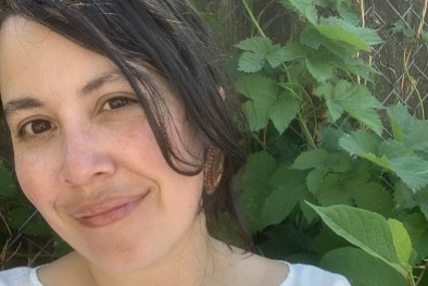 Close-up outdoor portrait of a person with dark hair wearing a white top, photographed in front of dense green foliage.
