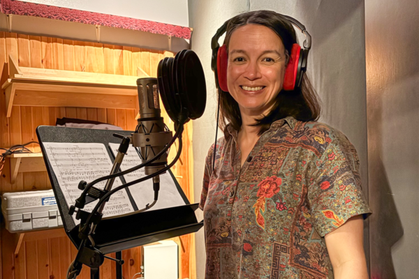 Person wearing red headphones stands at a microphone in a recording booth beside a music stand holding sheet music.
