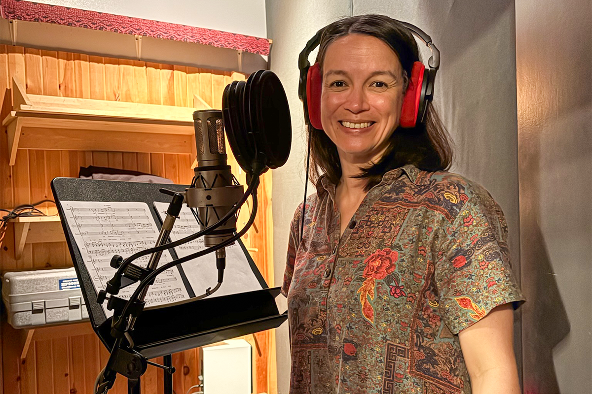 Person wearing red headphones stands at a microphone in a recording booth beside a music stand holding sheet music.
