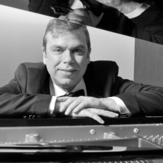 A black and white photo of a person wearing a tuxedo leaning against a piano.