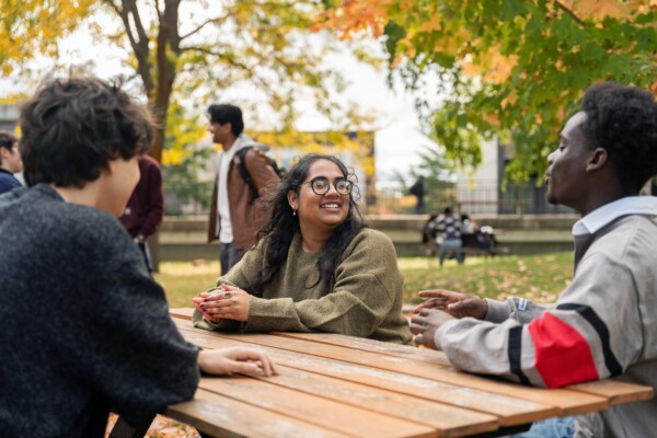 Several students sit and talk around a picnic table outdoors on a campus quad surrounded by autumn trees.