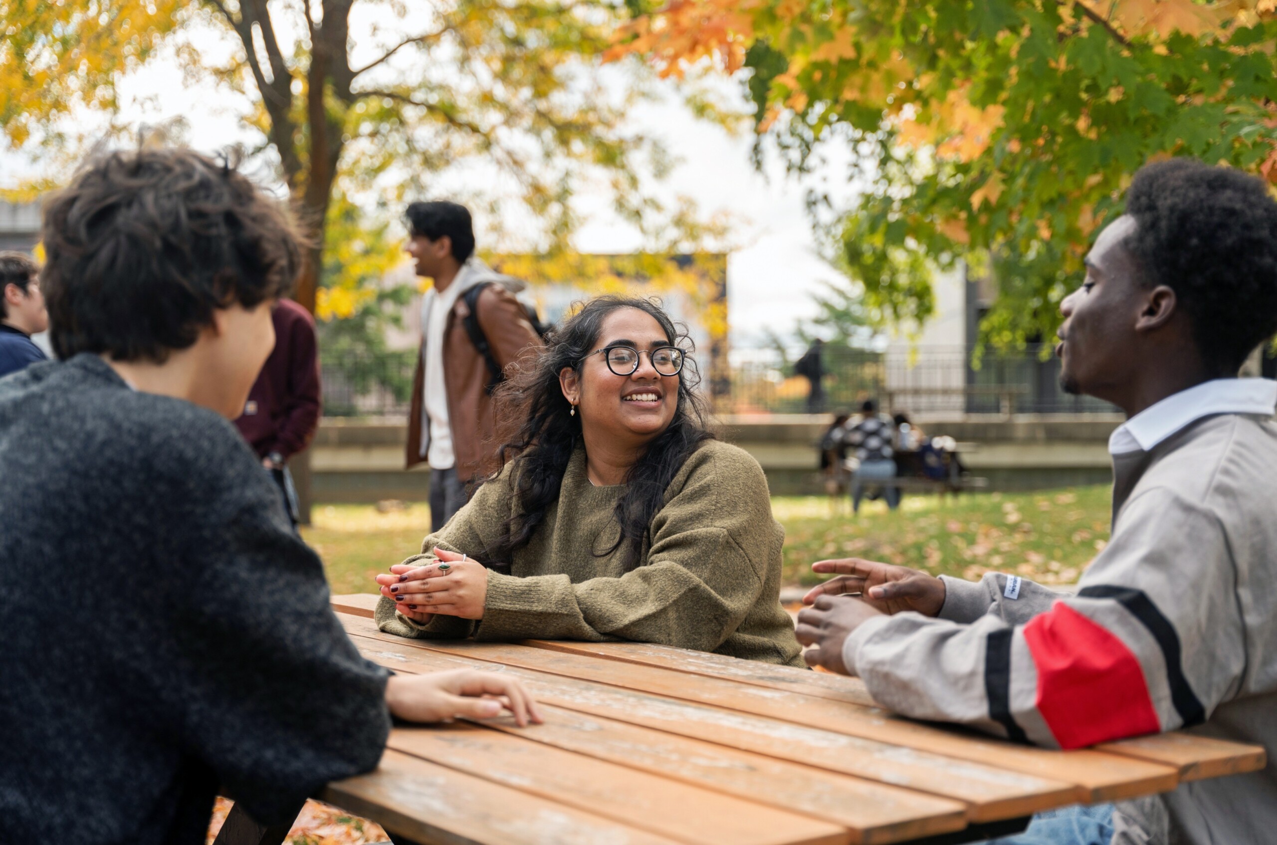 Several students sit and talk around a picnic table outdoors on a campus quad surrounded by autumn trees.