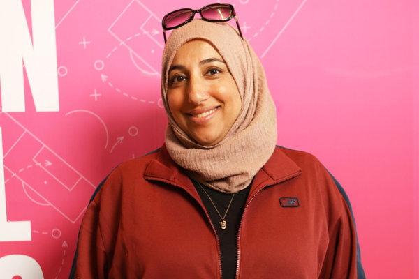 Person wearing a beige head scarf and a red zip-up jacket stands in front of a bright pink backdrop featuring white sports playbook-style graphics.