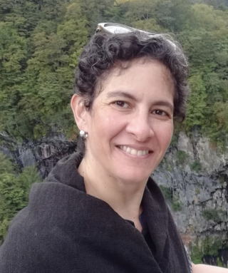 A person with short curly hair stands in front of a rocky cliff and forested landscape, with sunglasses on top of their head.