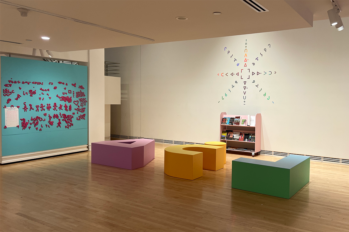 Three colourful chairs shaped like Cree letters in an open gallery space.