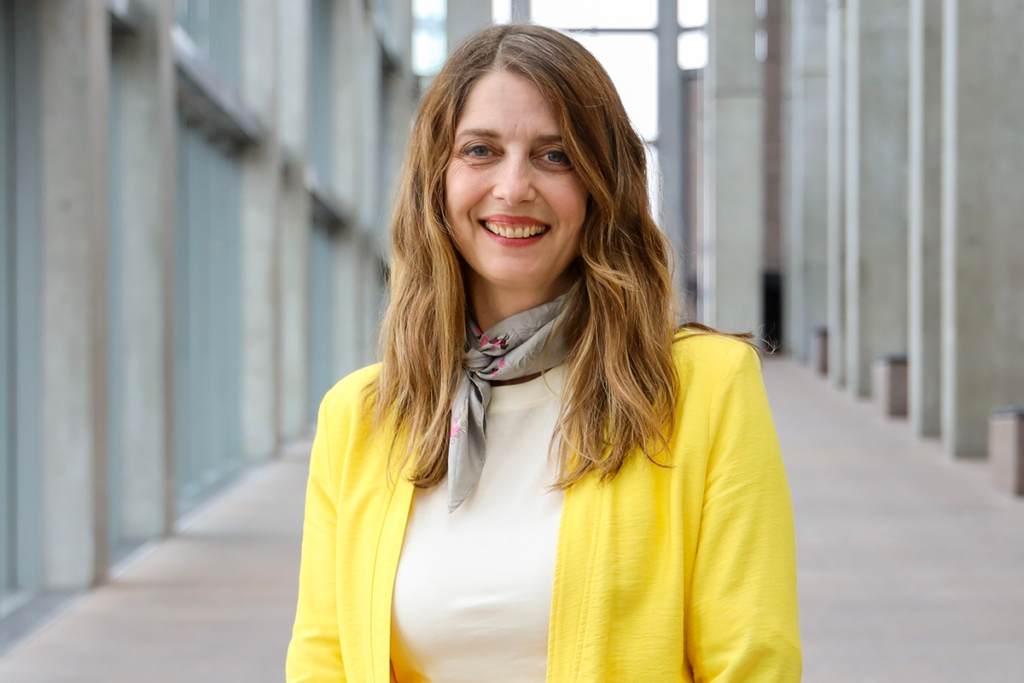 Catherine Sinclair, wearing a bright yellow blazer and patterned scarf, stands in a corridor with tall windows and columns.