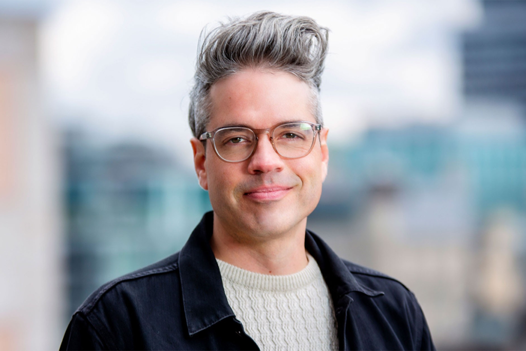 Jean-Noé Landry, wearing glasses, a light sweater and a dark jacket, stands in front of a softly blurred urban background.
