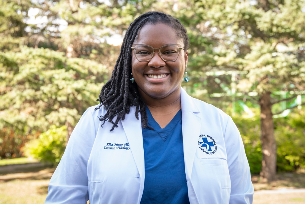 Kika Otiono, wearing a white medical coat over blue scrubs, stands outdoors in front of trees.