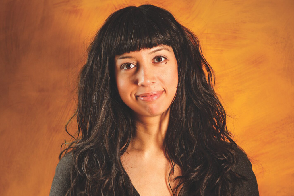 Saleema Nawaz, who has long dark hair and bangs, photographed against a warm orange backdrop.