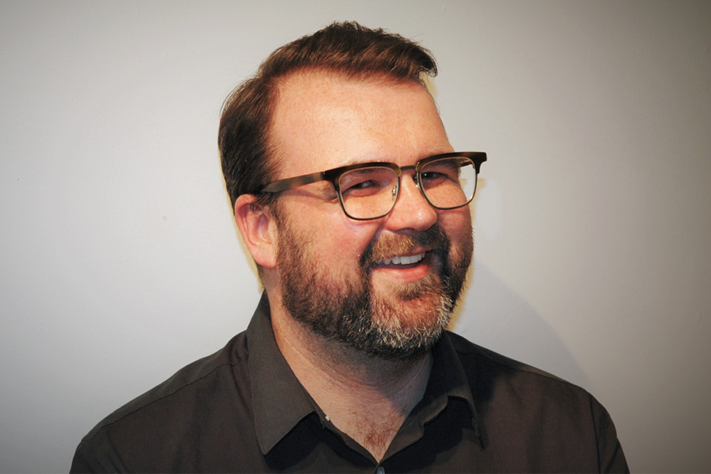 Sebastian Major, wearing glasses and a dark collared shirt, is photographed against a plain grey background.