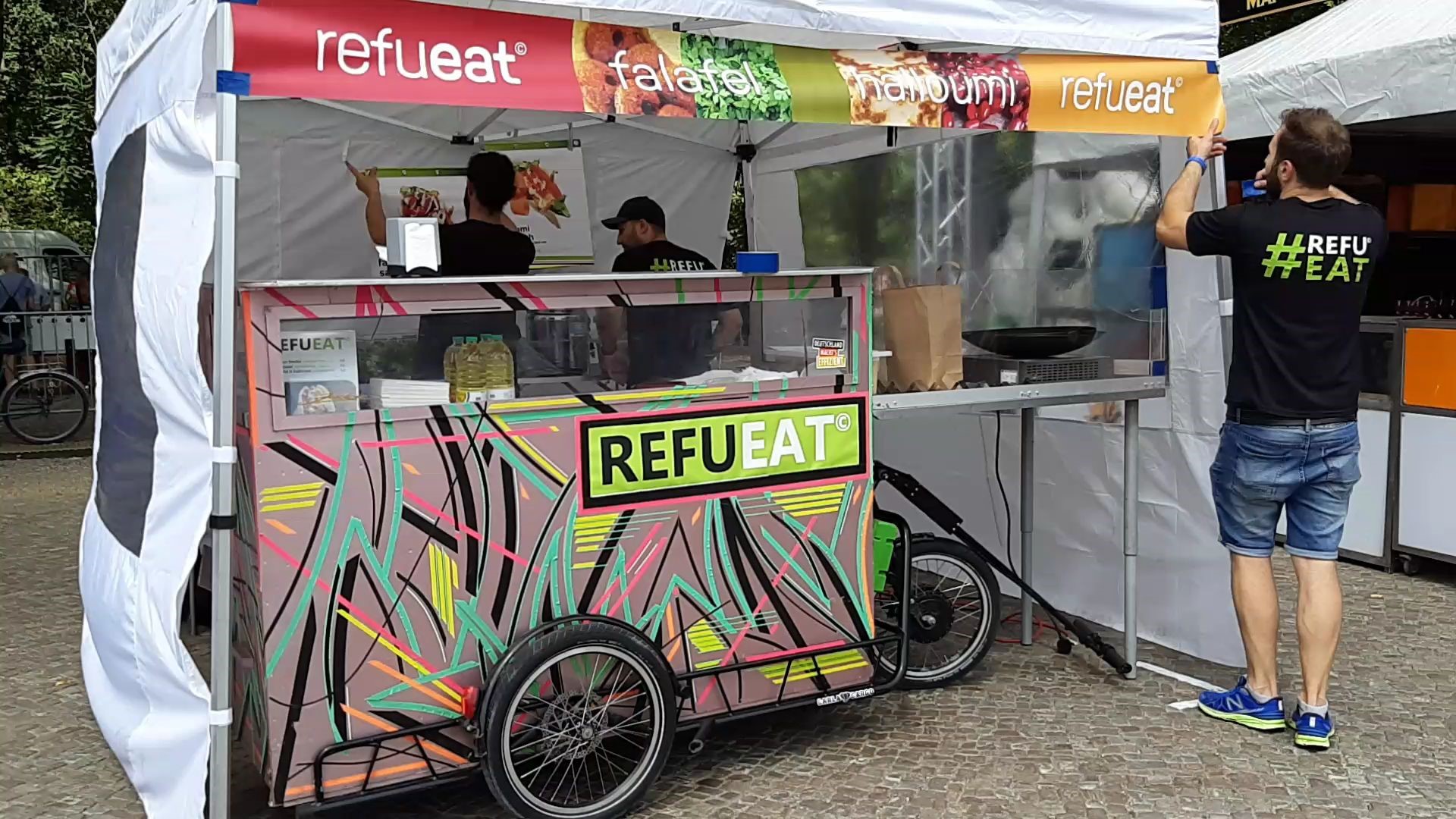 The RefuEat cart in a tent and preparing food for customers. The cart decorated with bright colours in abstract patterns.