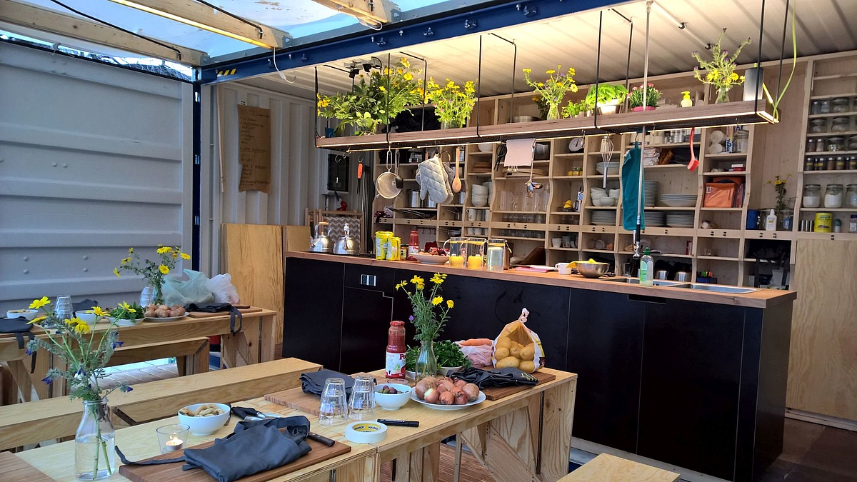 A full kitchen with wooden shelves and dining area with long wooden tables installed inside a shipping container.