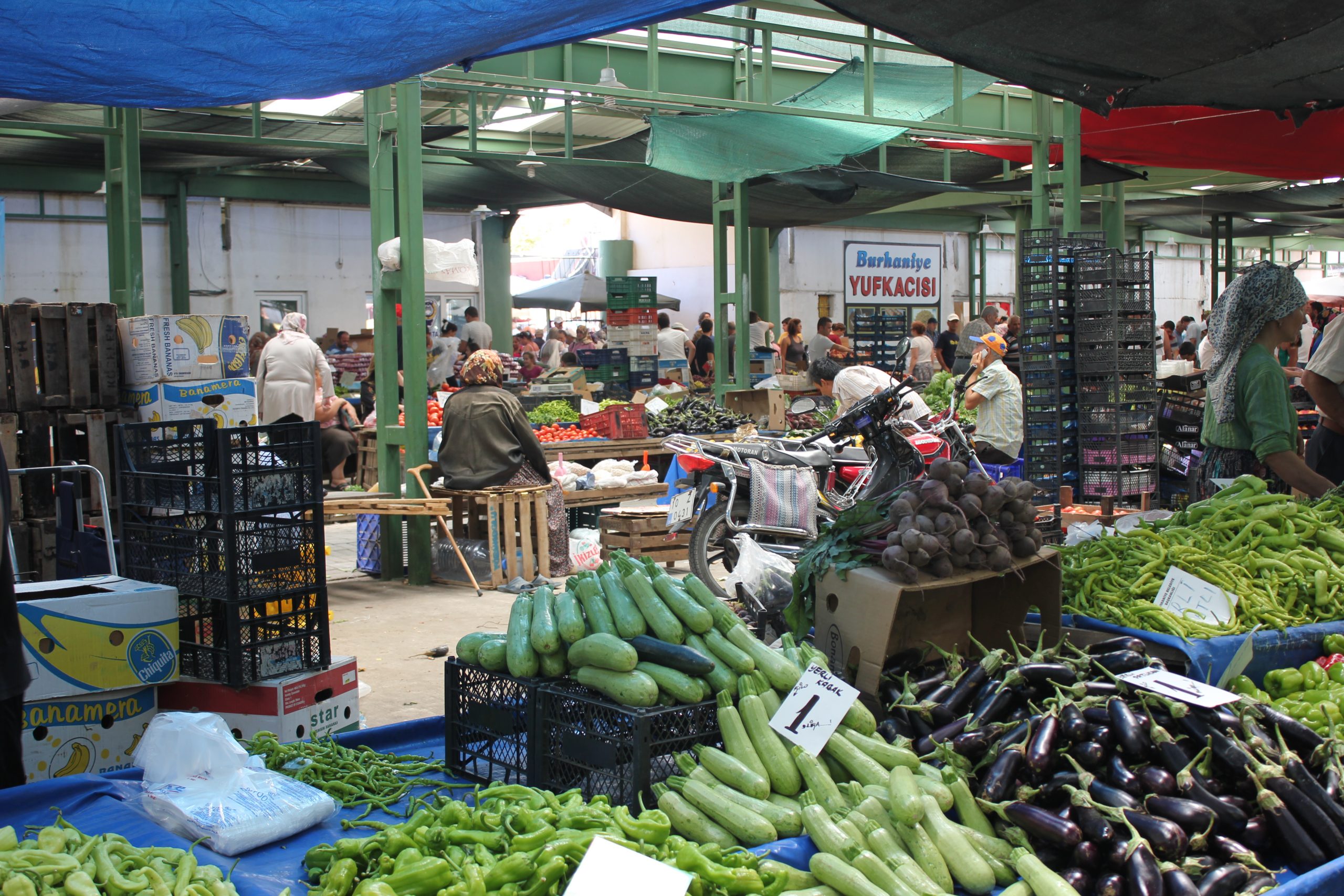 A number of people walk, sit, and shop under the tarp roofs of a vegtable market/bazaar in Burhaniye, Turkey.