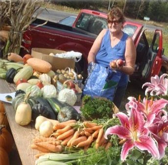 A woman behind a table of vegetables holding more produce in her hand. 