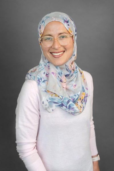 Amira Elghawaby smiles for a portrait photo against a dark grey background.