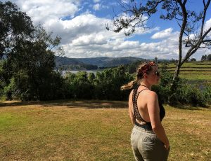 Kayley stands in a filed overlooking a view of mountains and water.