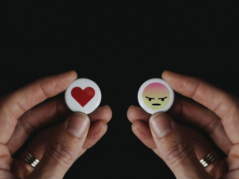 Hands holding a buttons, one with a heart icon and teh other an angry face.