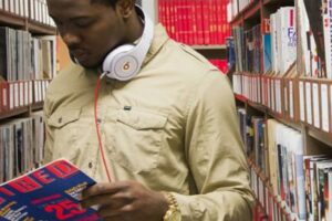 A student reads a magazine in the aisle of a library.