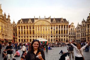 A street scene in Belgium in the forefront , a person holds a waffle while smiling at the camera