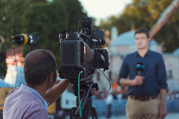 A camera operator features prominently in the foreground filming a reporter in the distance