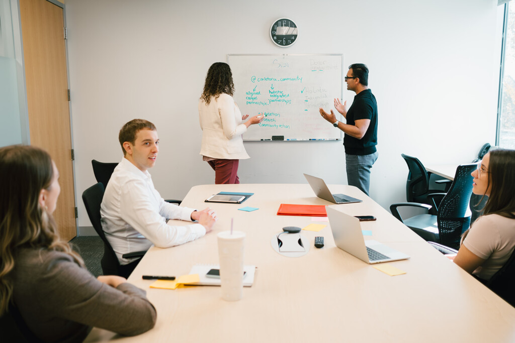 People work at a table in the foreground while two other people discuss at a whiteboard in the background.